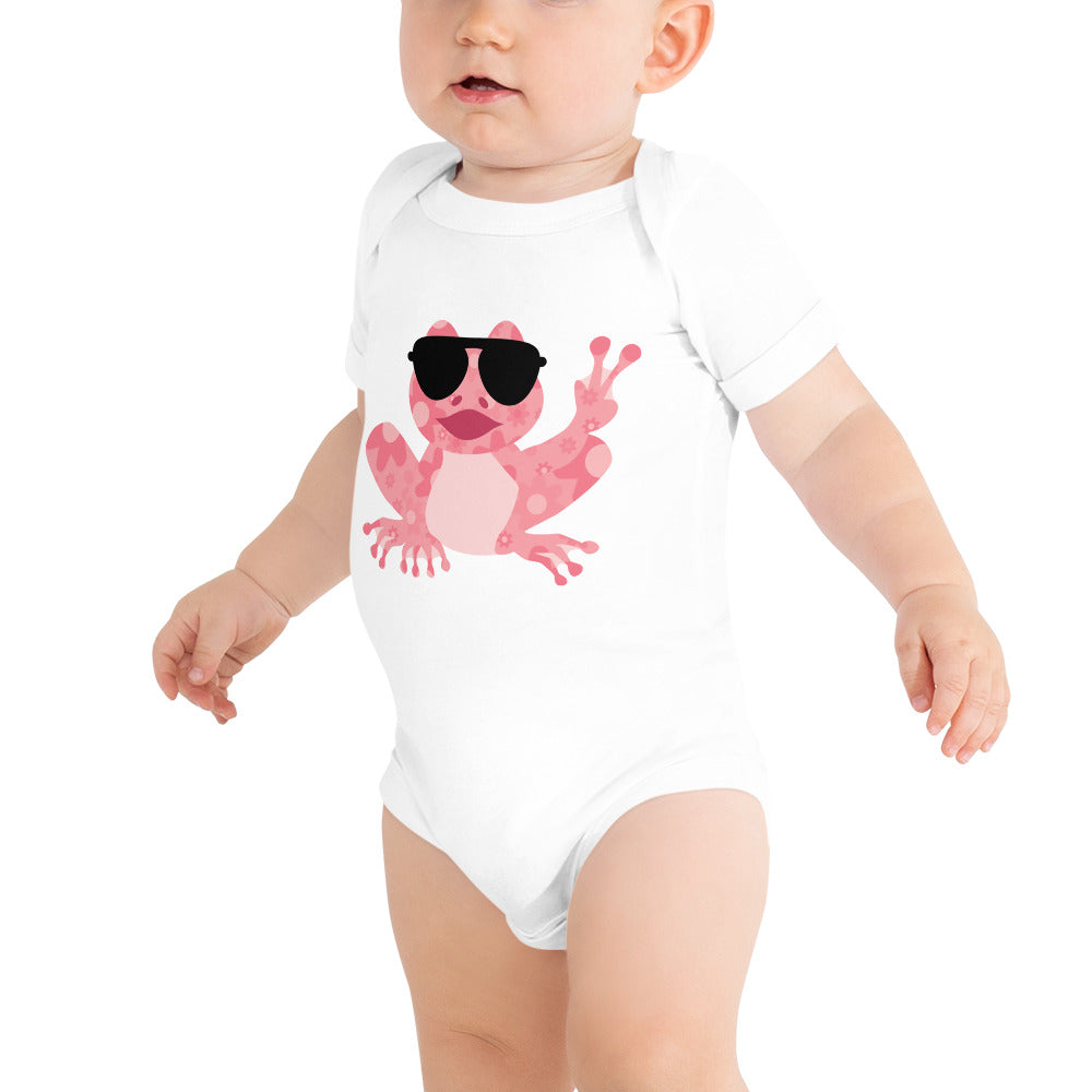 Shows a baby wearing the white one piece bodysuit with a pink frog with a floral pattern, wearing big black sunglasses and showing the peace sign, design is for baby girls